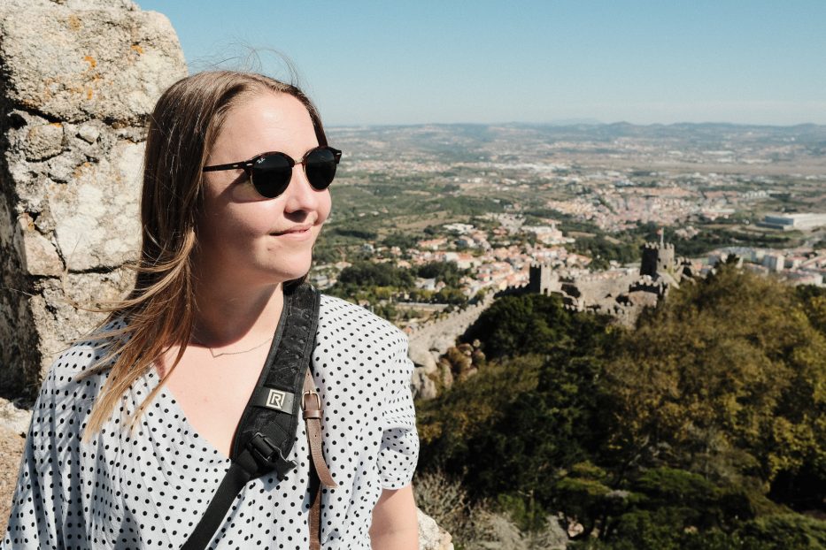 woman in a dotted dress with sunglasses looking out at a view point with Castelo dos Mouros in sintra in the background