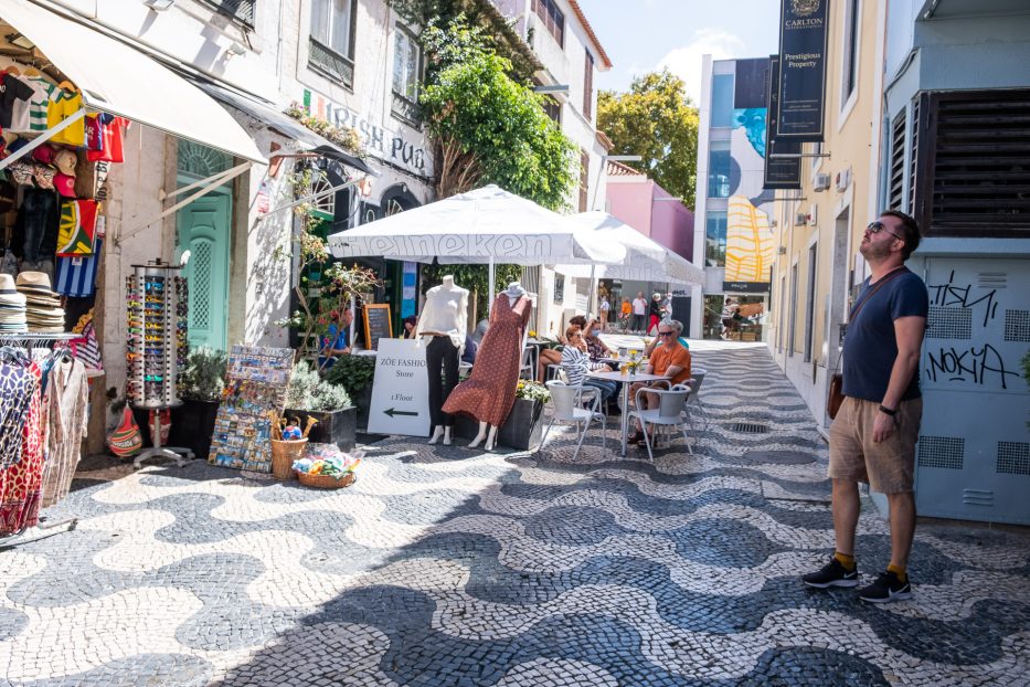man standing in a cobblestone street in Cascais with colorful building in the background