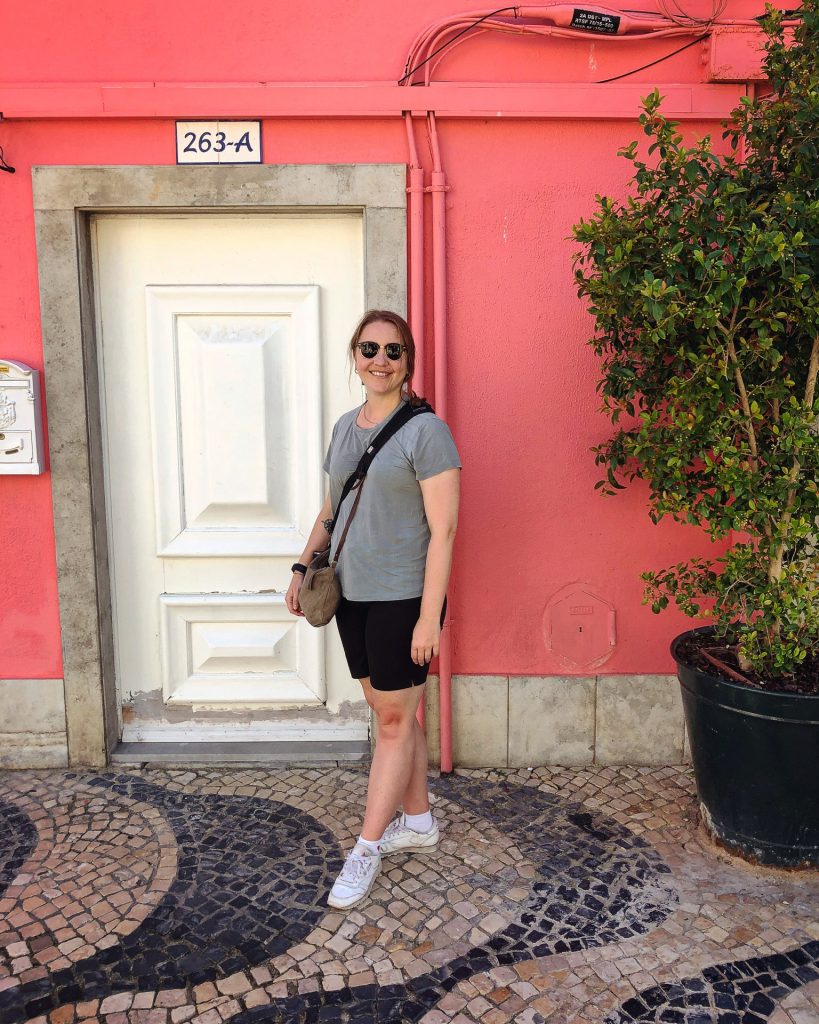 woman with sunglasses in front of a bright pink building and a door with the number 263-a