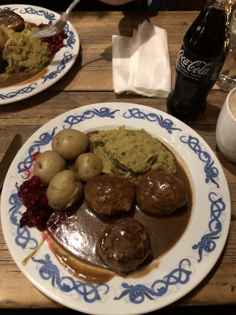 a plate filled with meat balls, potatoes and pea stew in a brown sauce and a bottle of Coca Cola on a wooden table