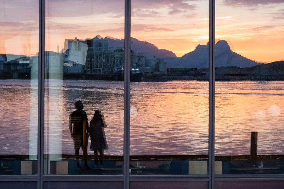 mann og kvinne står på kaia i Bodø med refleksjon av havet og fjell og solnedgang i et vindu