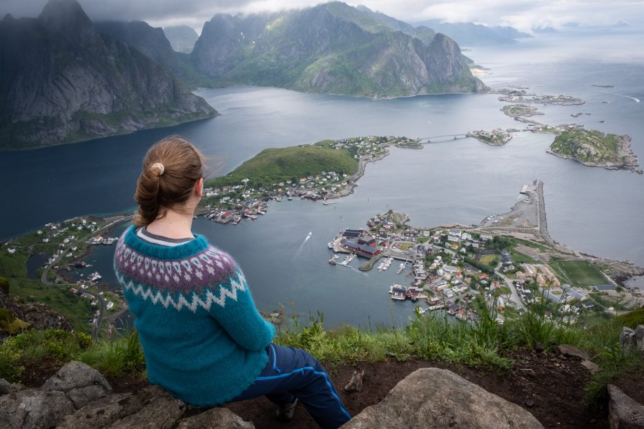 woman in a colorful knitted sweater sitting on top of reinebringen looking down at reine in Lofoten norway