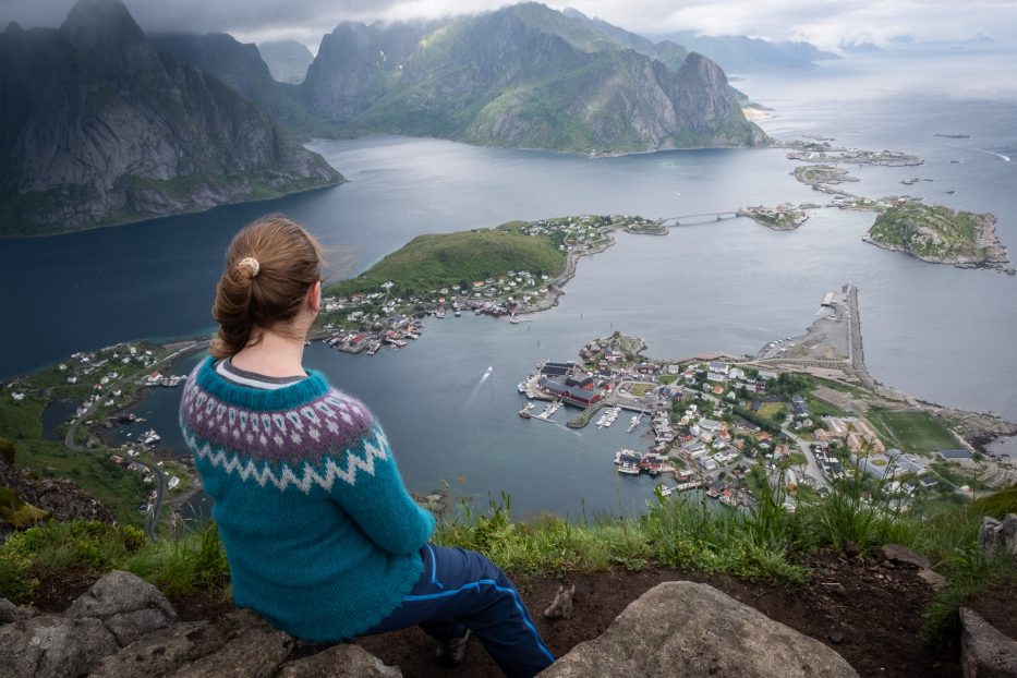 woman in a colorful knitted sweater sitting on top of reinebringen looking down at reine in Lofoten norway