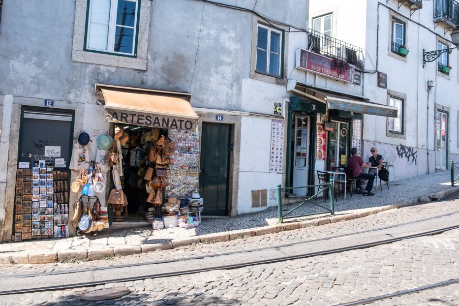 mennesker sitter utenfor en kafé og en liten butikk i en bratt bakke i Alfama