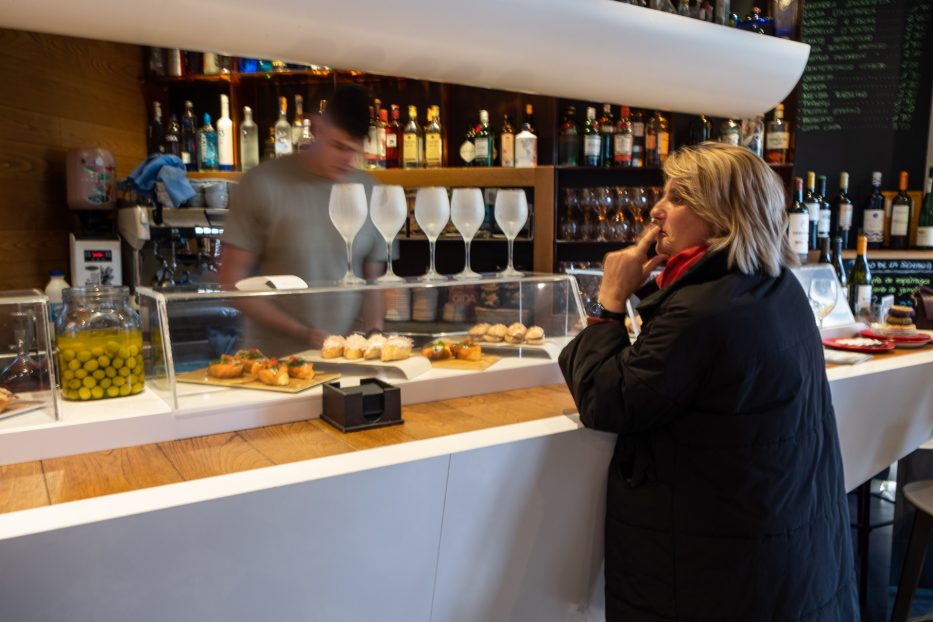 woman in front of Pintxos bar with five glasses of txakoli on top and a man behind working