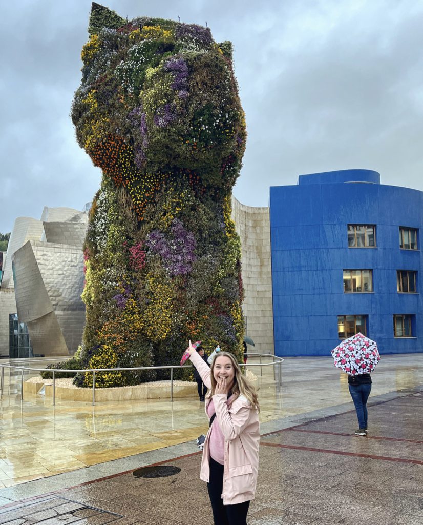 statuen puppy utenfor guggenheimmuseet i bilbao og en kvinne i rosa regnjakke peker på den og ser glad ut