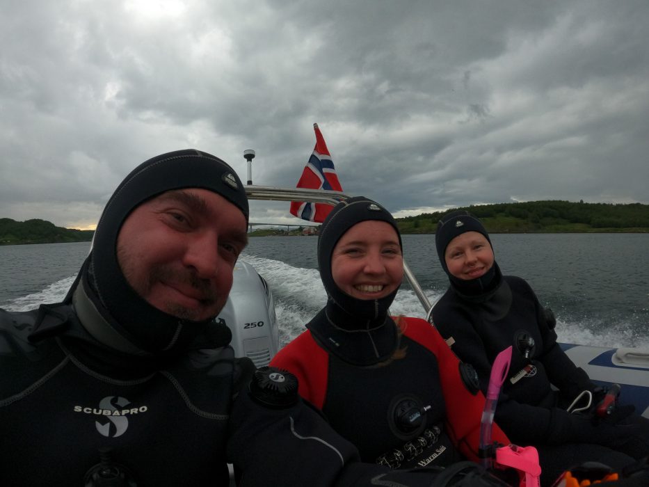 three people with dry suits on in a boat with a Norwegian flag