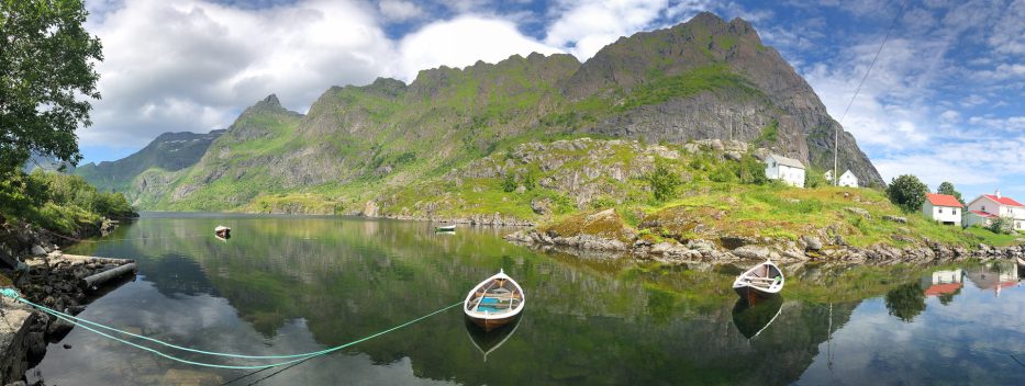 a panorama of a calm lake with row boats and houses and a big mountain in lofoten norway