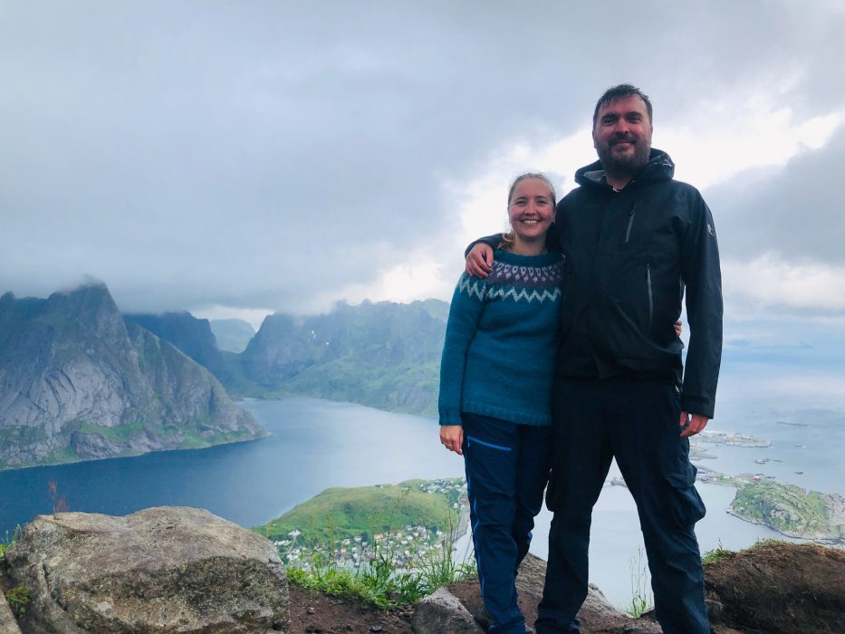 kvinne og mann står og smiler til fotografen på toppen av reinebringen med utsikten bak seg