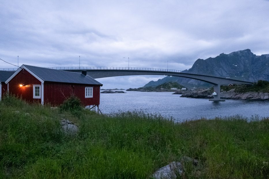 en rød rorbu og en bro i bakgrunnen med fjell i bakkant
