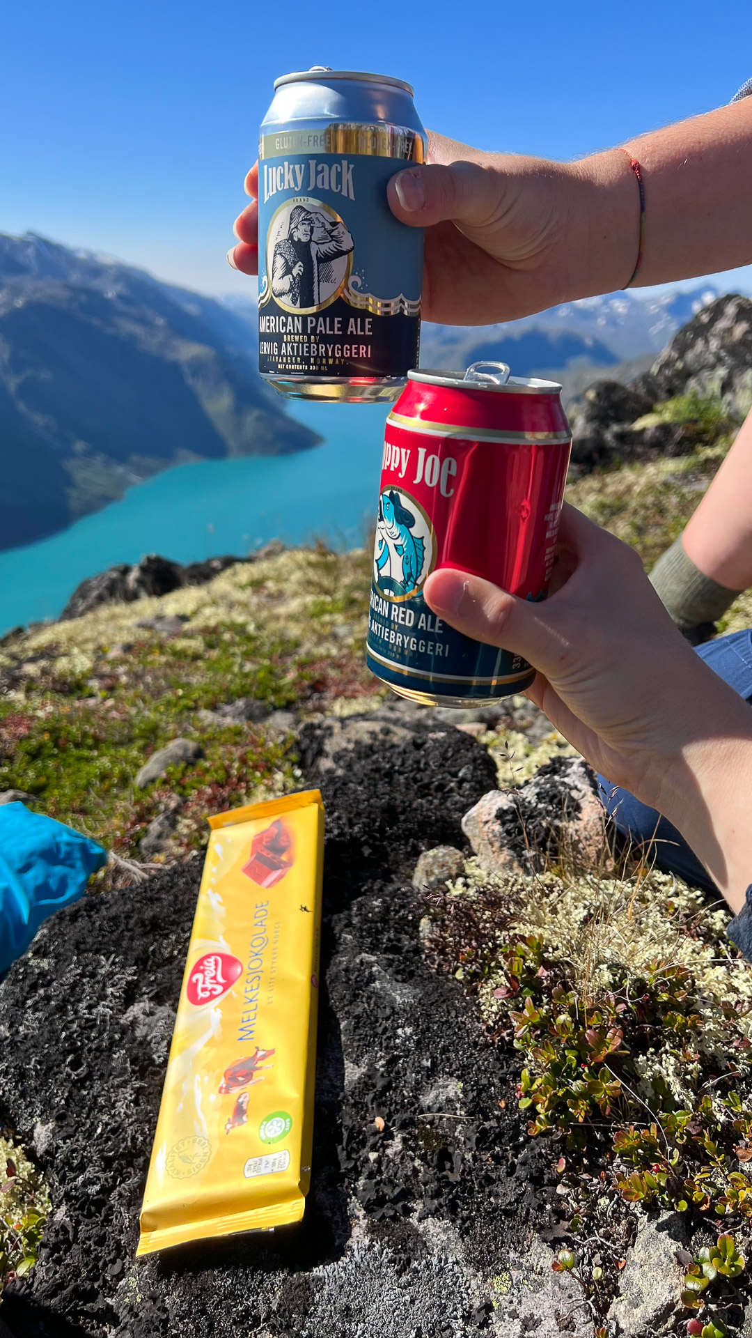 norsk melkesjokolade og to øl holdt opp foran nydelig utsikt ved besseggen