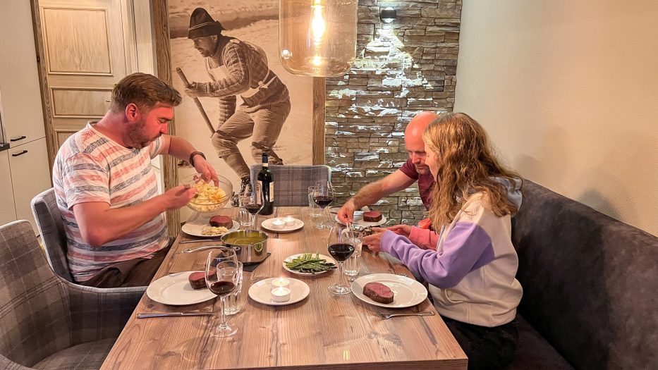 two men and a woman eating steaks inside a cabin at beitostølen