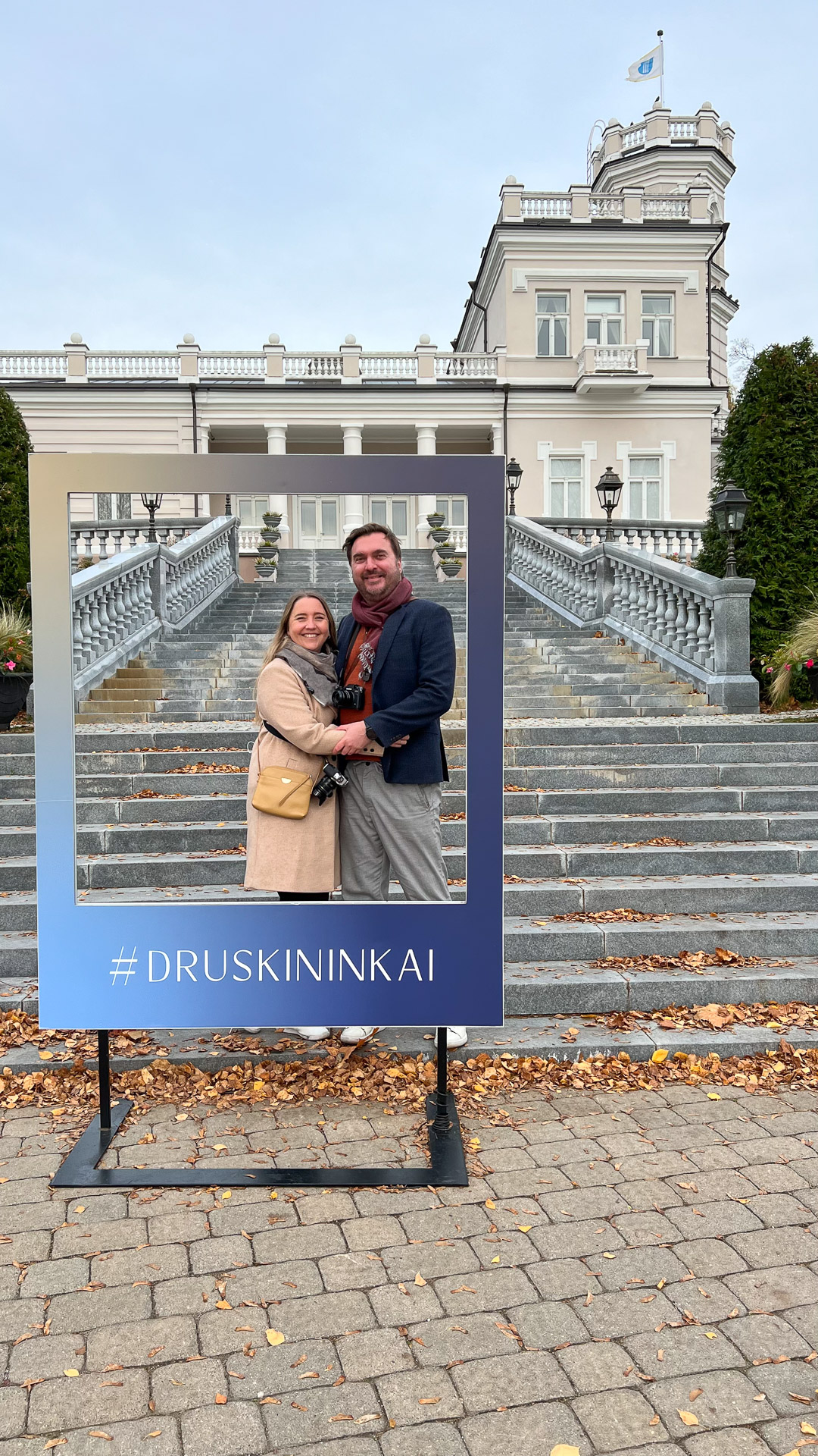 man and woman standing inside a frame with #druskininkai written on it in front of famous house