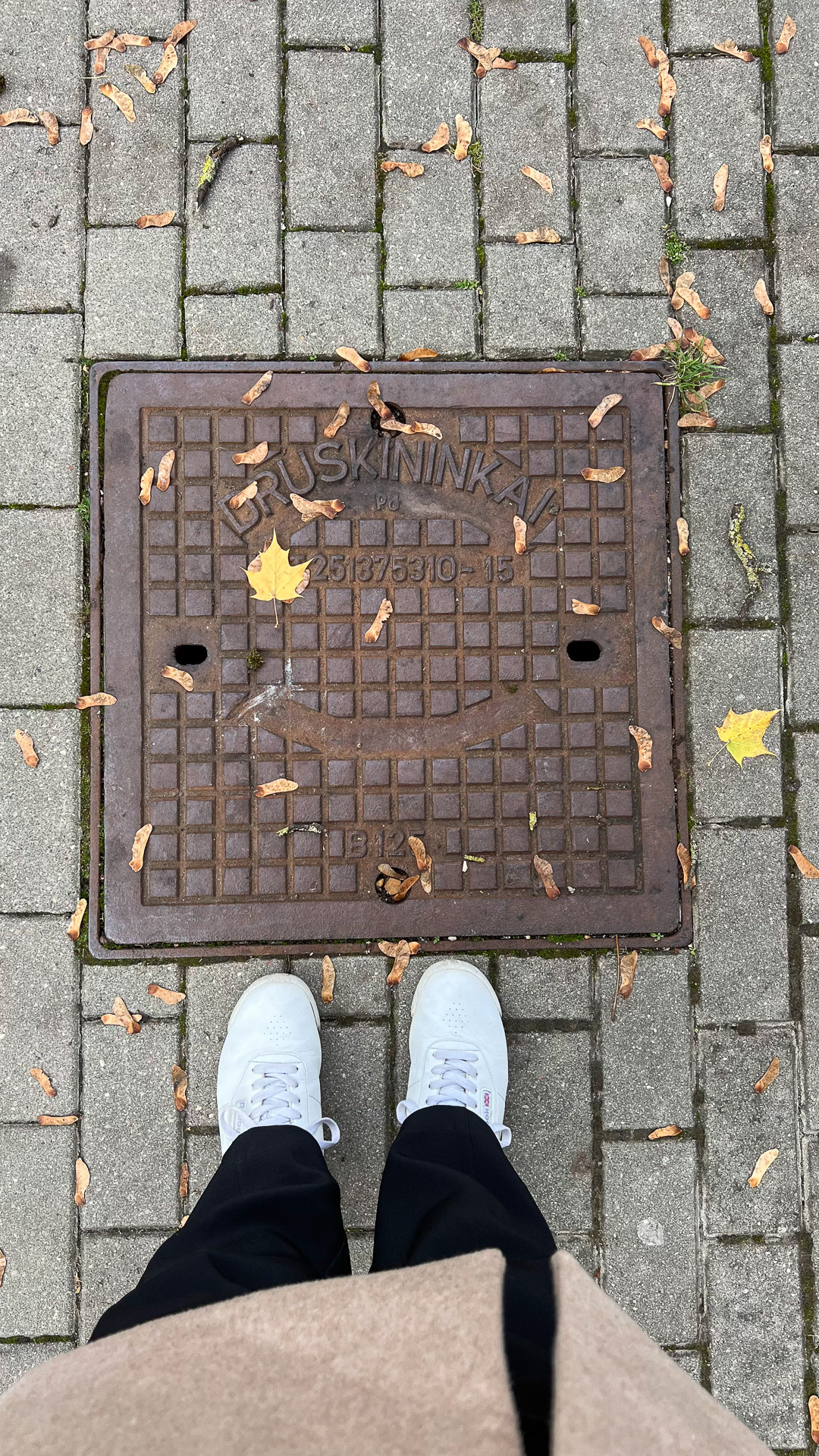 person with brown jacket black pants white reebok shoes standing over a manhole cover saying druskininkai