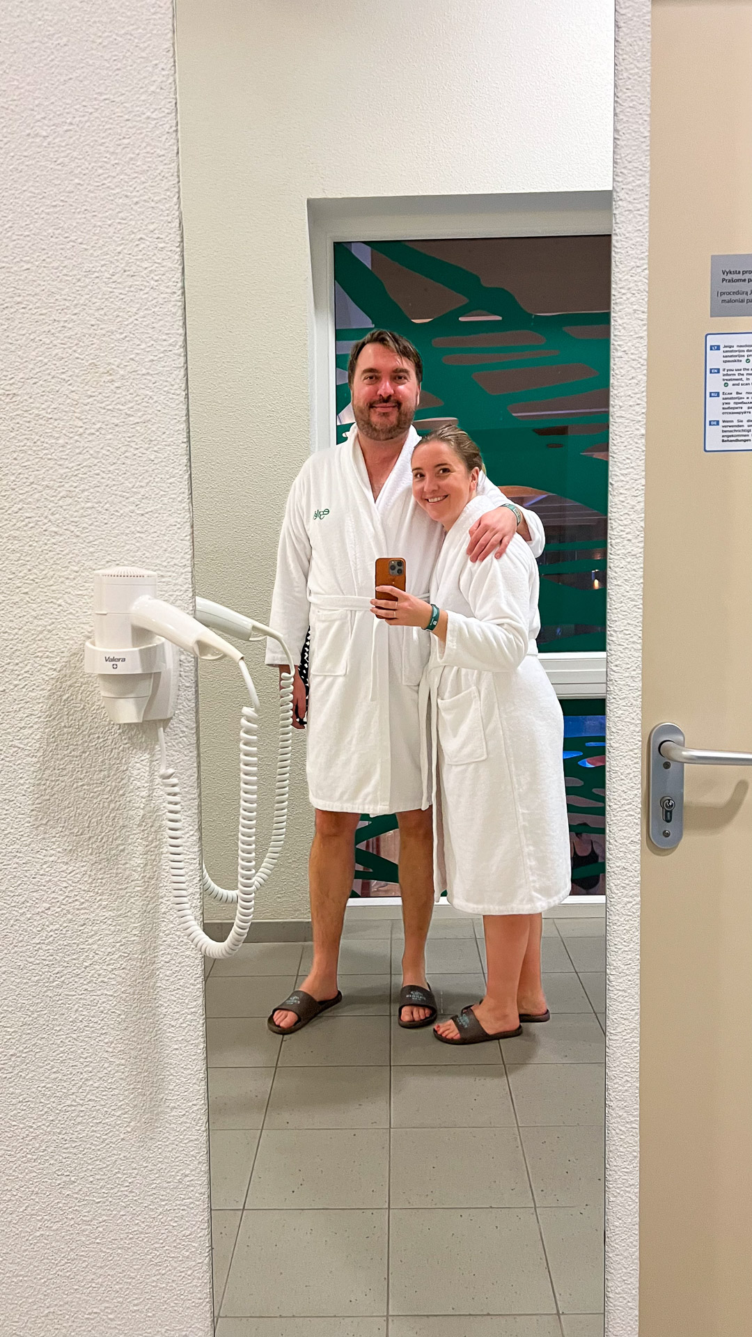 man and woman wearing white bathrobes taking a selfie in a mirror