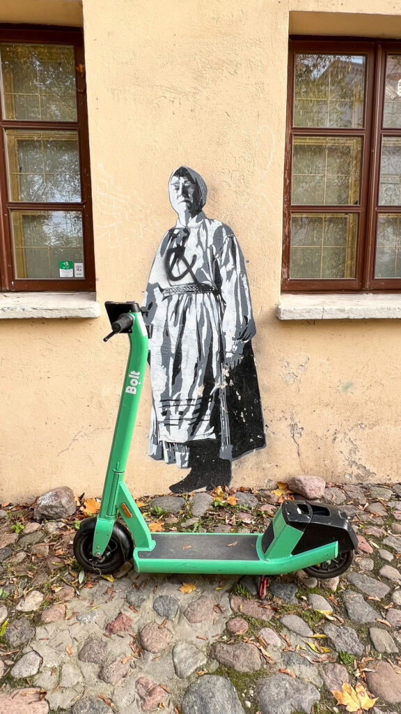 gatekunst av en gammel kone i tradisjonelt litauisk antrekk og en grønn el-sparkesykkel fra bolt foran