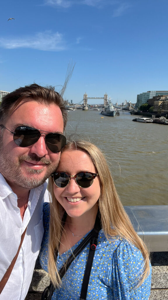 man and woman smiling while wearing sunglasses on london bridge with tower bridge in the background