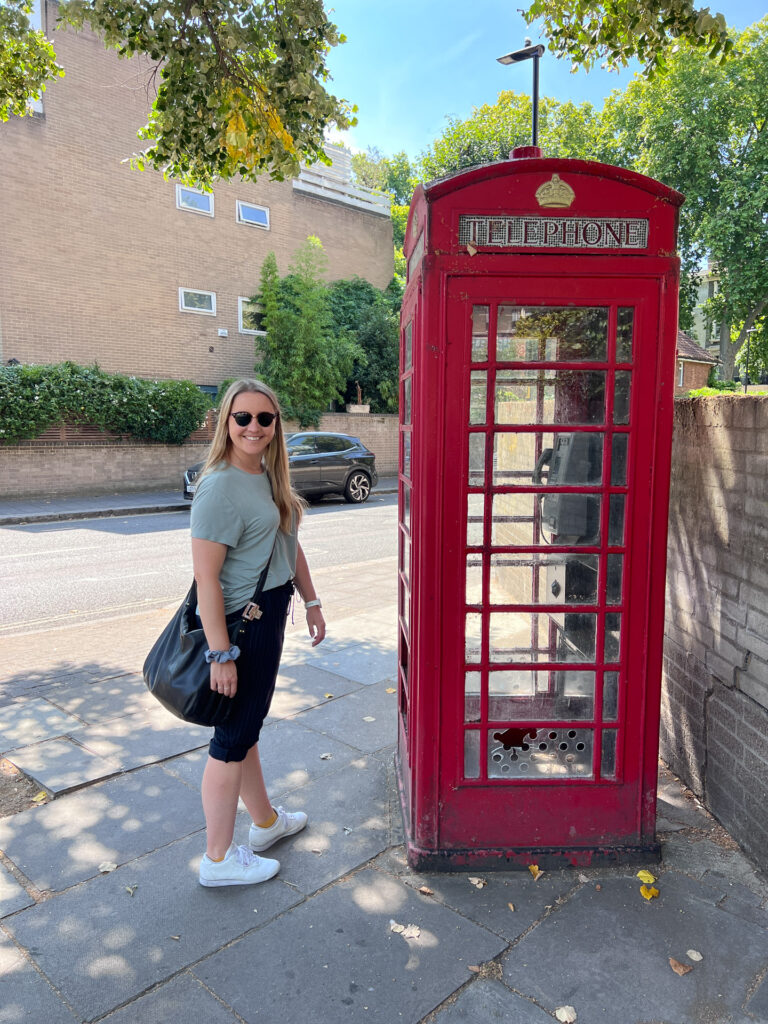 smilende kvinne poserer utenfor rød telefonkiosk i london