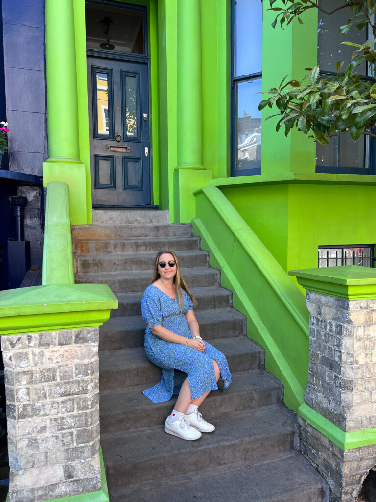 kvinne i blå kjole med solbriller sitter i trapp utenfor knall grønt hus med blå dør i notting hill london