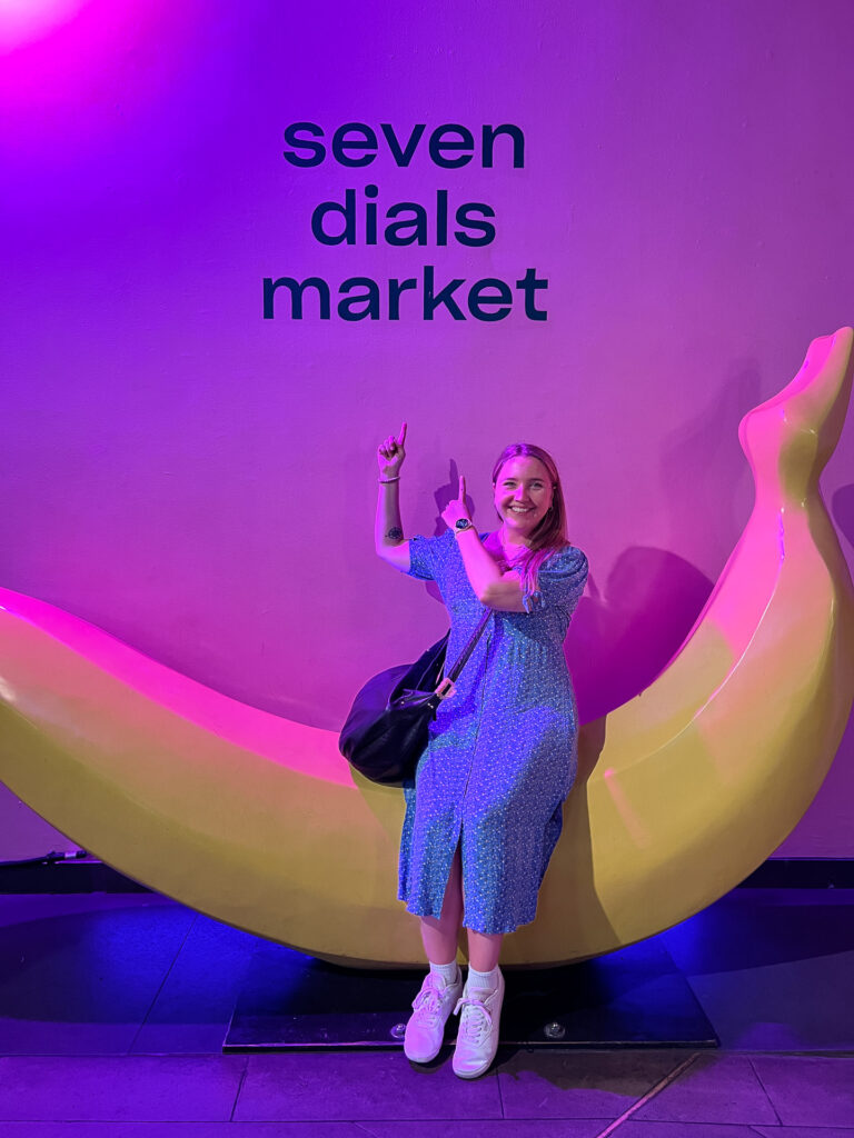 smilende kvinne i blå kjole sitter på gul banan og peker på sorte bokstaver på vegg som sier seven dials market