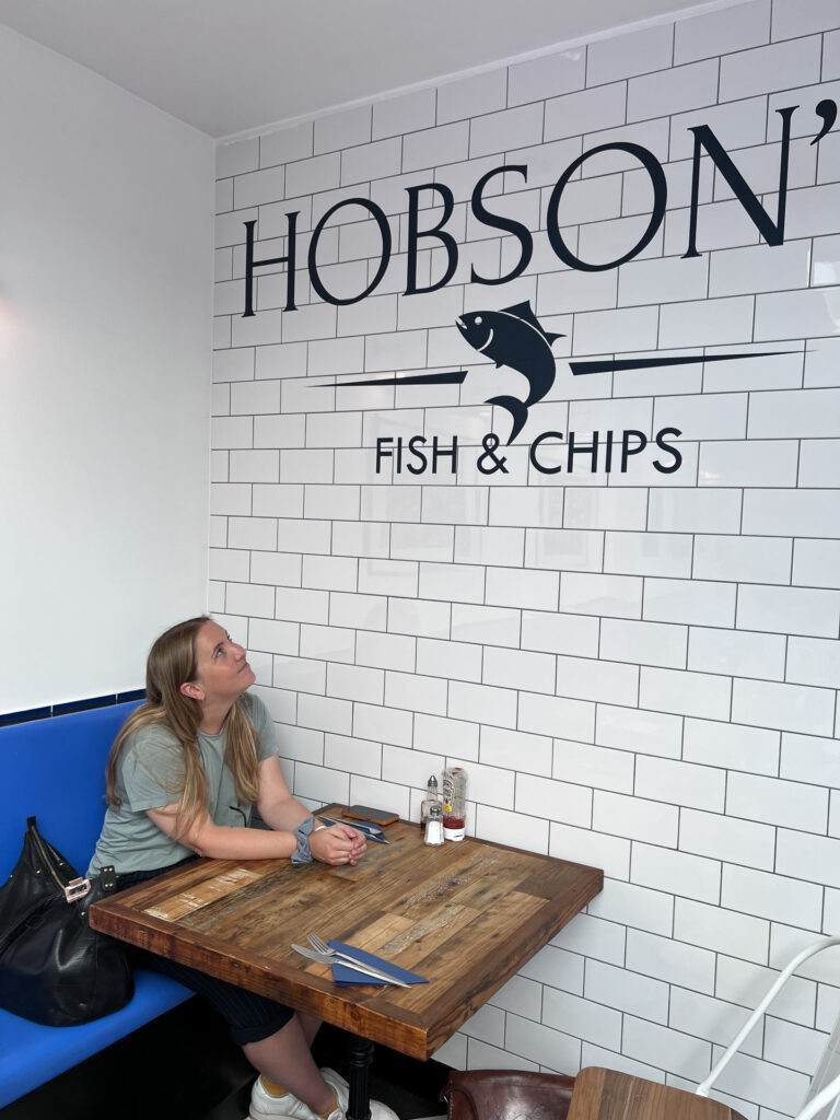 en kvinne sitter med hendene på bordet mens hun ser på veggen der man ser logoen til hobson's fish & chips i london