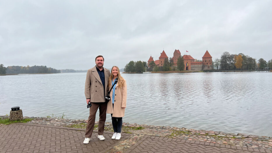 smiling man and woman standing in front of the island with trakai castle