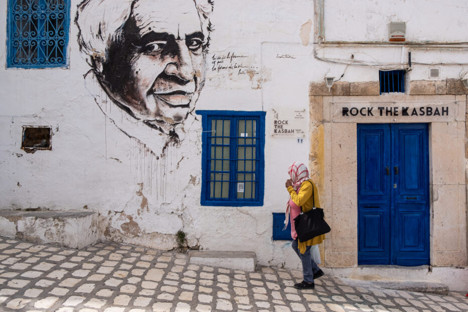 a woman with yellow jacket and pink top with a scarf on her head walks past rock the kasbah and street art in sidi bou said