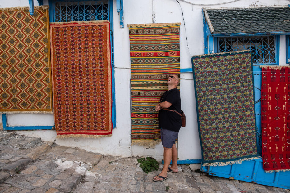 a man laughs in front of colourful carpets on a white house with blue details in sidi bou said