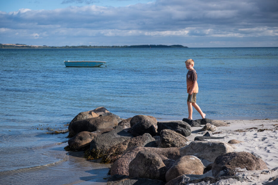 a boy walking on the beach in front of a small boat in the water