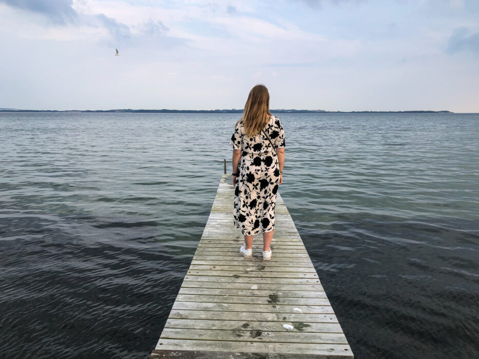 a woman in a long dress facing away from the camera standing on a floating bridge in the water