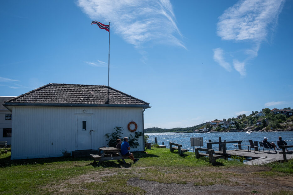 people enjoying the sun next to glomma river on a pier and in front of a white house with a norwegian flag on top