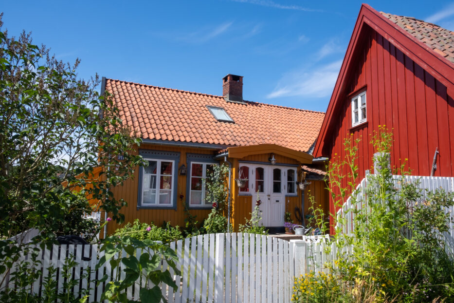 a yellow and a red house stands side by side with a white fence and green plants in front