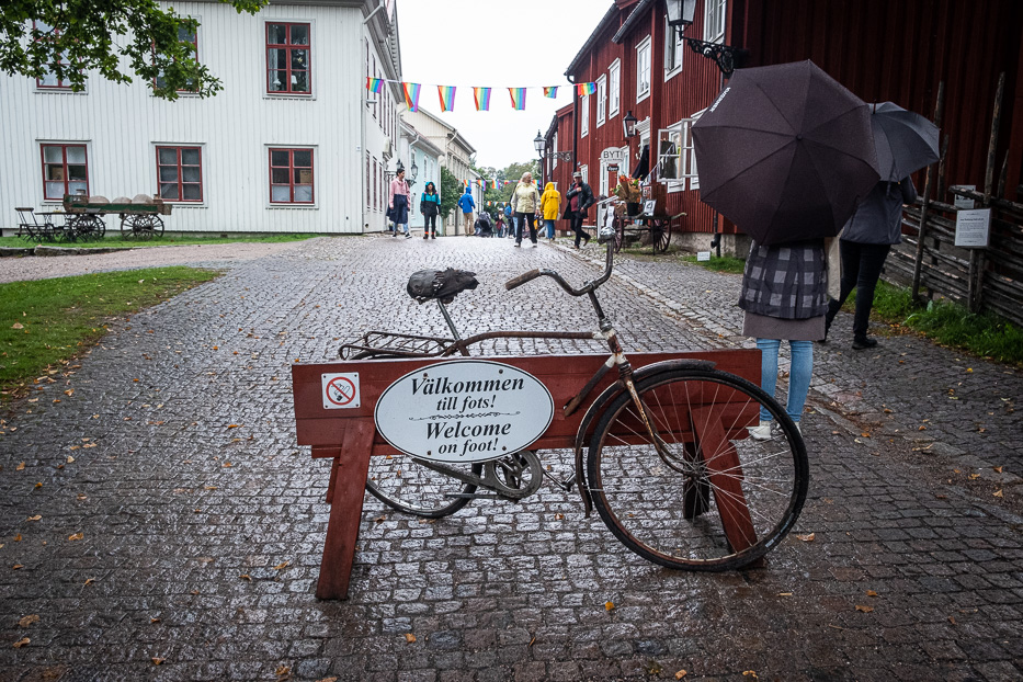 person with umbrella and a bicycle in front of sign saying welcome on foot in wadköping area sweden
