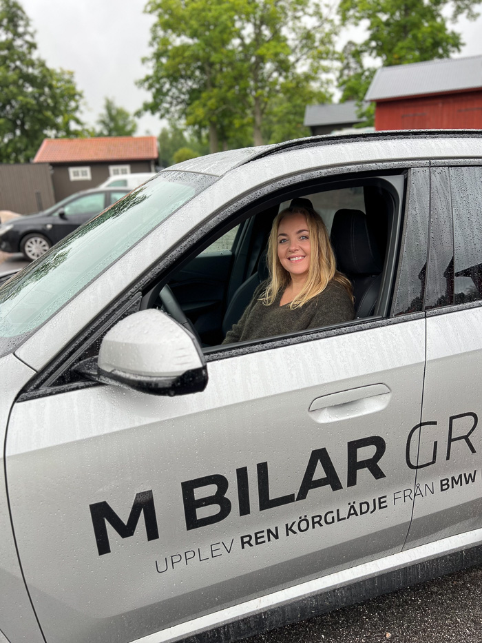 en blond kvinne smiler utenfor en grå bil med teksten m bilar group på siden parkert utenfor Katrinelund Gästgiveri & Sjökrog