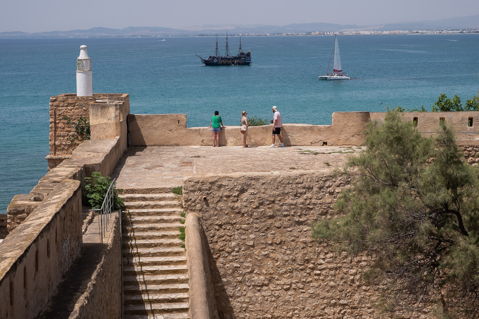 three people standing on the top of hammamet fortress looking out at a pirate ship and a sail boat at sea